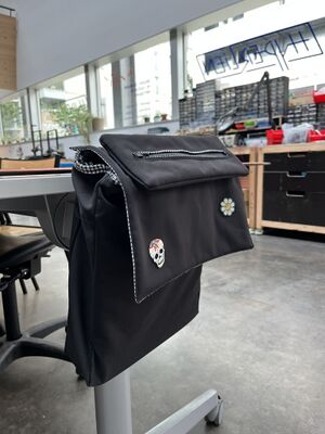 Sacoche rectangulaire avec abattant et trousse extra-plate scratchée sur l'abattant. Le sac est accroché à une table comme sur un accoudoir de fauteuil roulant.